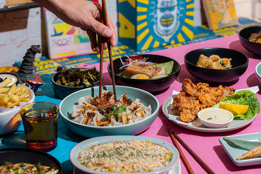 A close-up of a hand using wooden chopsticks to pick up dumplings from a light blue bowl on a vibrant pink table covered with various colorful Japanese dishes, including fried chicken, miso Mac and cheese, and side salads.