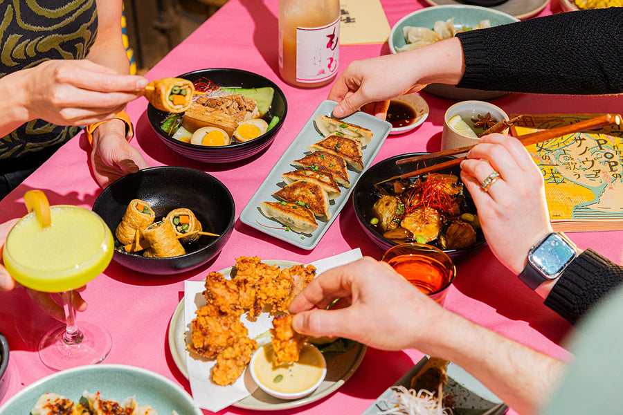 A close-up, overhead view of a vibrant pink restaurant table at Chotto Motto, laden with an assortment of Japanese dishes, including gyoza,  chicken karaage, sashimi, and cocktails, with multiple hands reaching in to grab food.