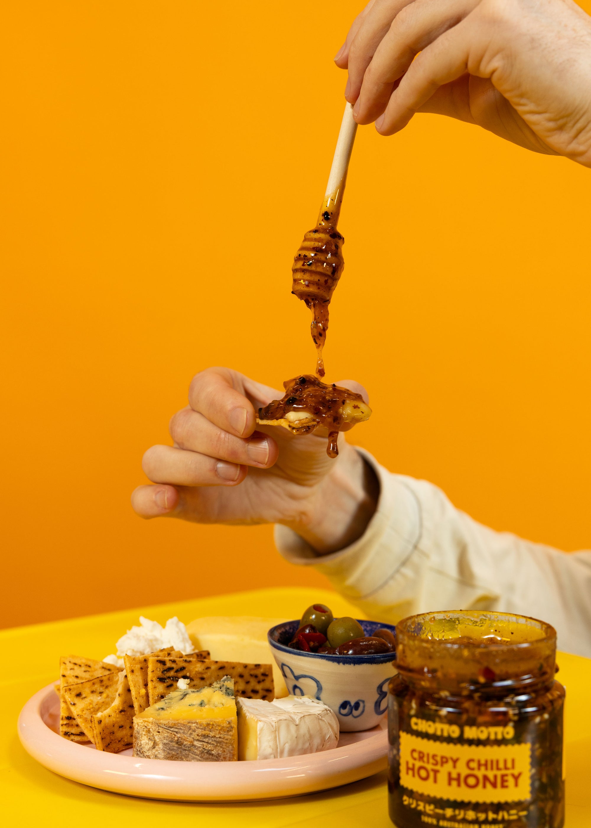 Person drizzling  Crispy Chilli Hot Honey from a jar onto a plate of cheese against an orange background