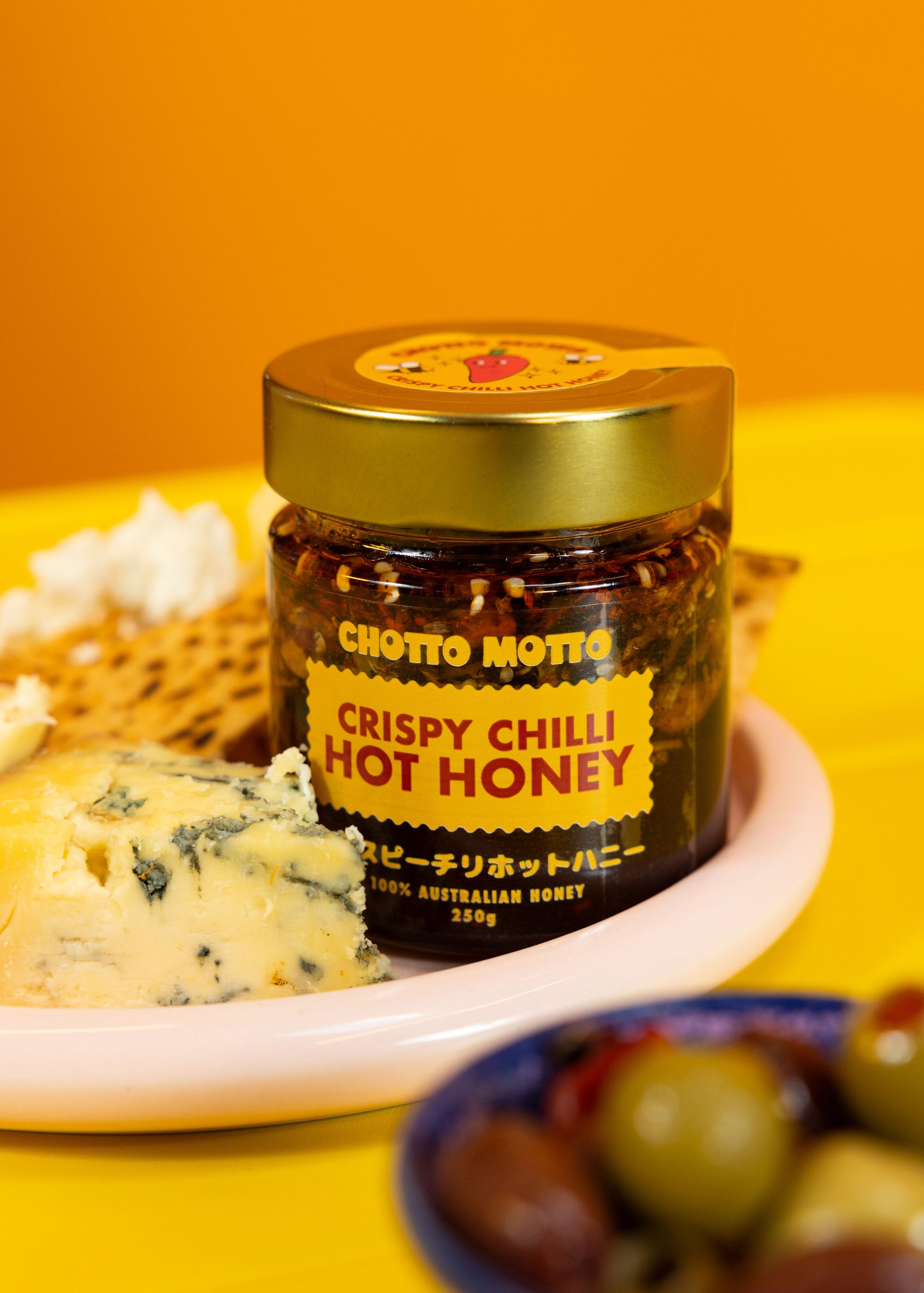 Jar of Chotto Motto Crispy Chilli Hot Honey on a plate with cheese and olives against an orange background